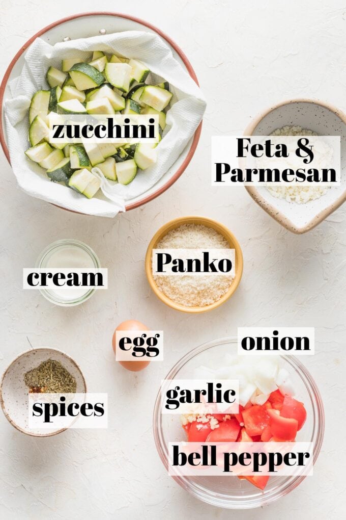 Labeled overhead photo of chopped zucchini and bell pepper, onion, minced garlic, feta and Parmesan cheese, egg, heavy cream, salt, black pepper, red pepper, and Italian seasoning measured and arranged in prep bowls.