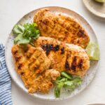Overhead view of three chicken breasts marinated in a Baja style resting on a plate garnished with lime wedges and fresh cilantro.