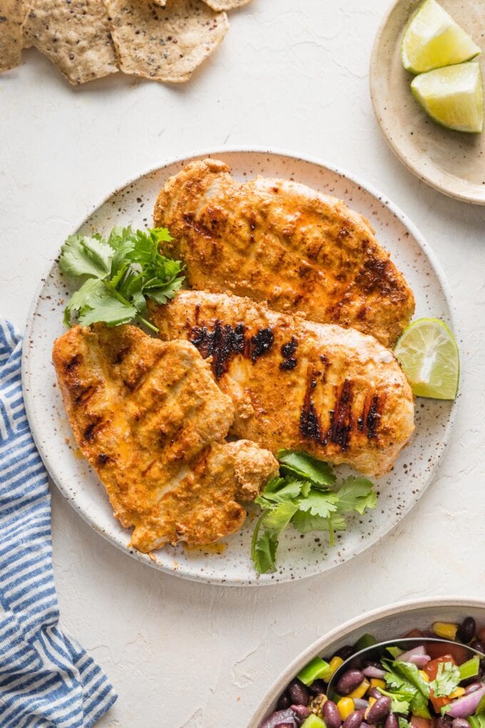 Overhead view of three chicken breasts marinated in a Baja style resting on a plate garnished with lime wedges and fresh cilantro.