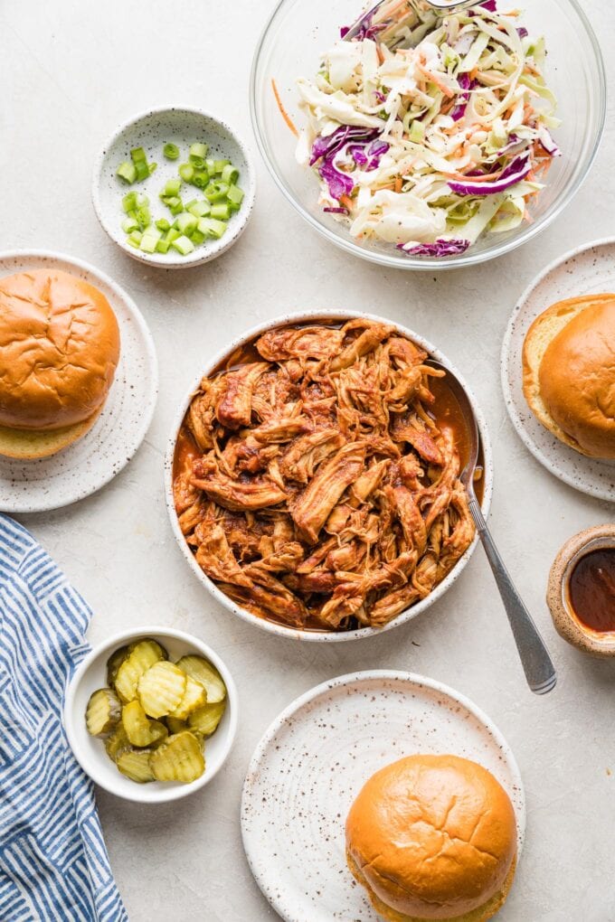 Overhead image showing a bowl of pulled BBQ chicken, another bowl of coleslaw, toasted brioche buns, pickles, and sliced green onions.