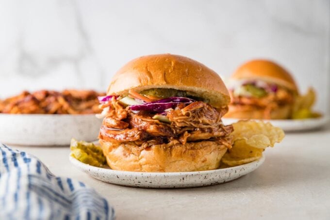 Landscape image of a BBQ chicken sandwich served on a small plate with pickles and chips.