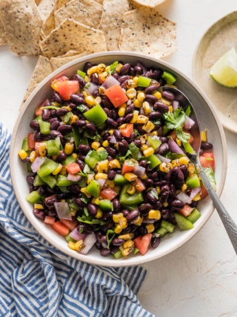 Serving bowl full of black bean salsa surrounded by tortilla chips and extra lime wedges.