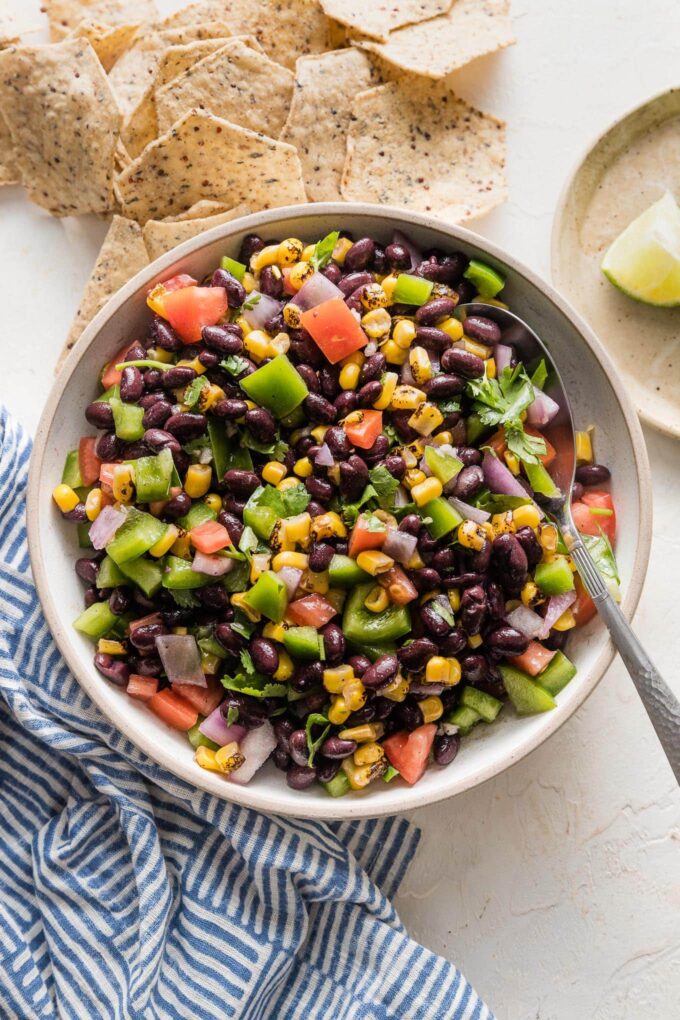 Serving bowl full of black bean salsa surrounded by tortilla chips and extra lime wedges.