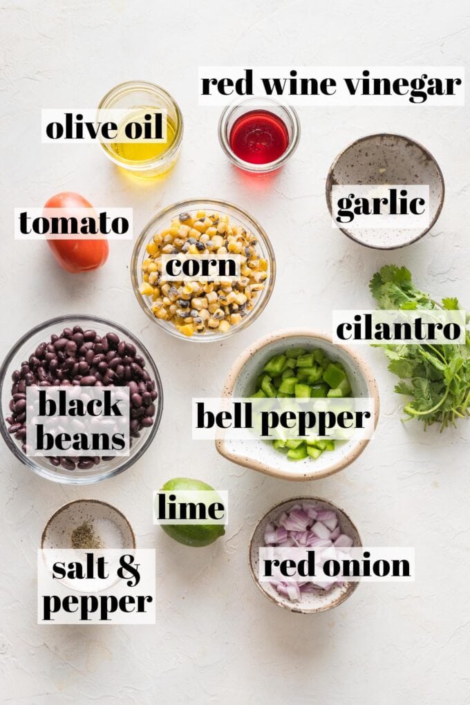 Labeled overhead view of chopped red onion, green bell pepper, corn kernels, minced garlic, drained and rinsed black beans, cilantro leaves, olive oil, red wine vinegar, a lime, salt, and black pepper arranged in prep bowls.
