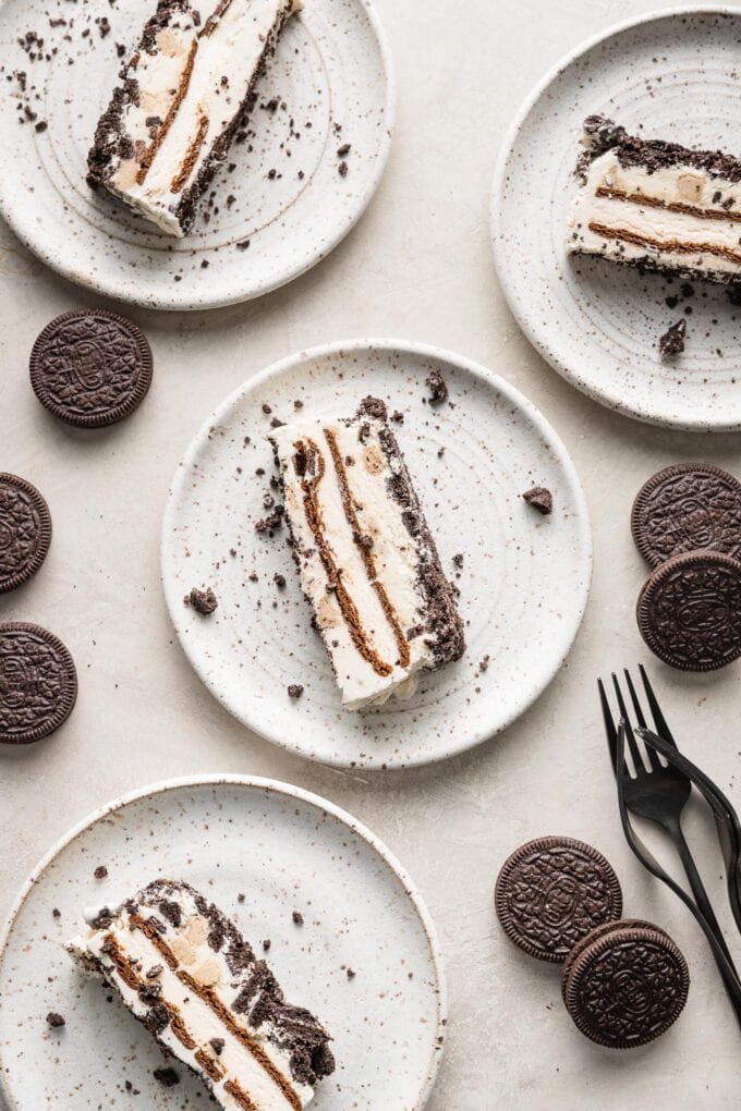 Set of four dessert plates with a slice of Oreo ice cream cake on each, with forks and extra Oreos scattered on the table nearby.