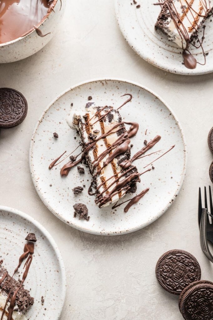 Close up of one slice of Oreo ice cream cake on a dessert plate, with a bowl of melted chocolate to the side ready to drizzle on top.