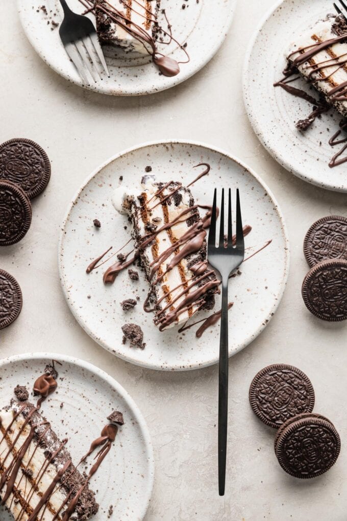 Set of four dessert plates with a slice of Oreo ice cream cake on each, with forks and extra Oreos scattered on the table nearby.