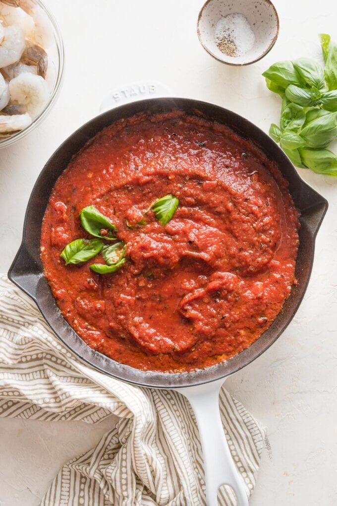 Skillet full of homemade marinara sauce with fresh basil and raw shrimp in the background waiting to be added.