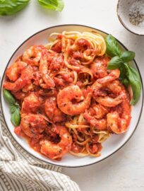 Small white plate piled high with pasta and shrimp cooked in a homemade marinara sauce, garnished with fresh basil leaves.