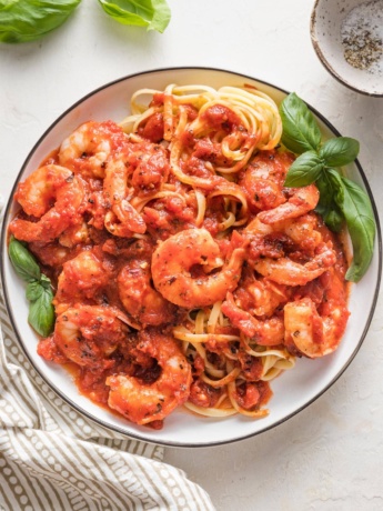 Small white plate piled high with pasta and shrimp cooked in a homemade marinara sauce, garnished with fresh basil leaves.