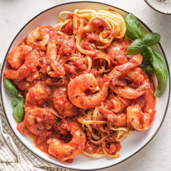 Small white plate piled high with pasta and shrimp cooked in a homemade marinara sauce, garnished with fresh basil leaves.