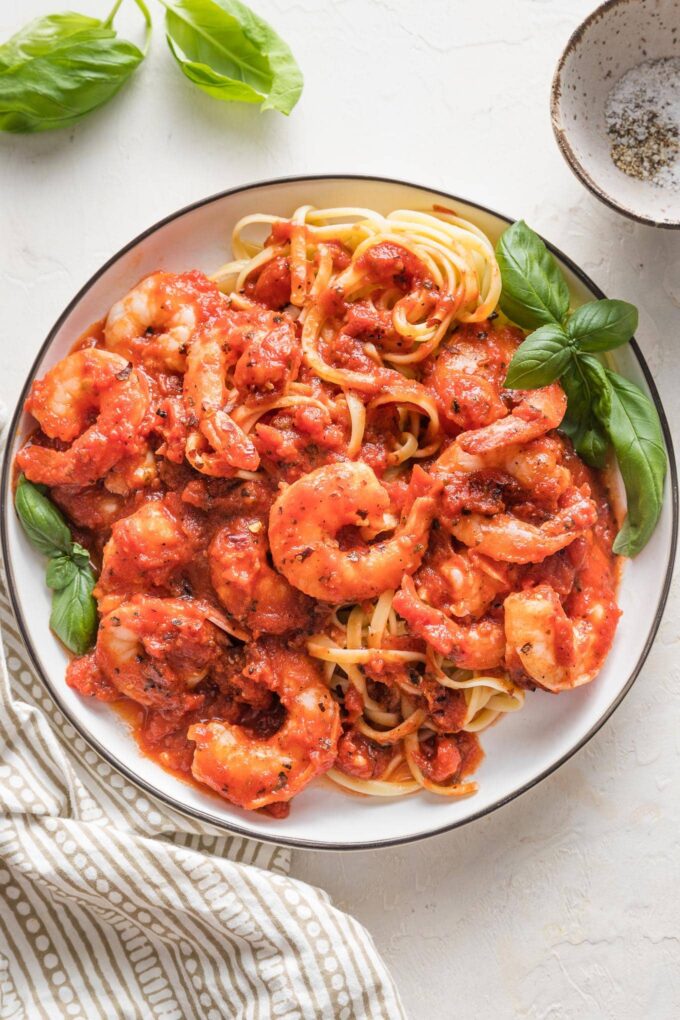 Small white plate piled high with pasta and shrimp cooked in a homemade marinara sauce, garnished with fresh basil leaves.