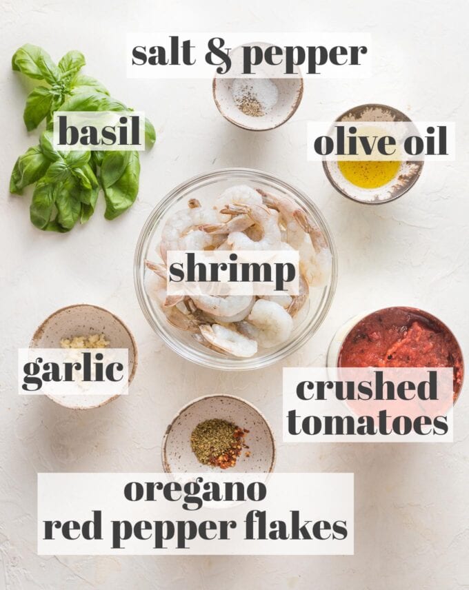 Labeled overhead photo of raw shrimp, crushed tomatoes, minced garlic, olive oil, fresh basil leaves, dried oregano, and red pepper flakes measured into prep bowls and ready to cook.