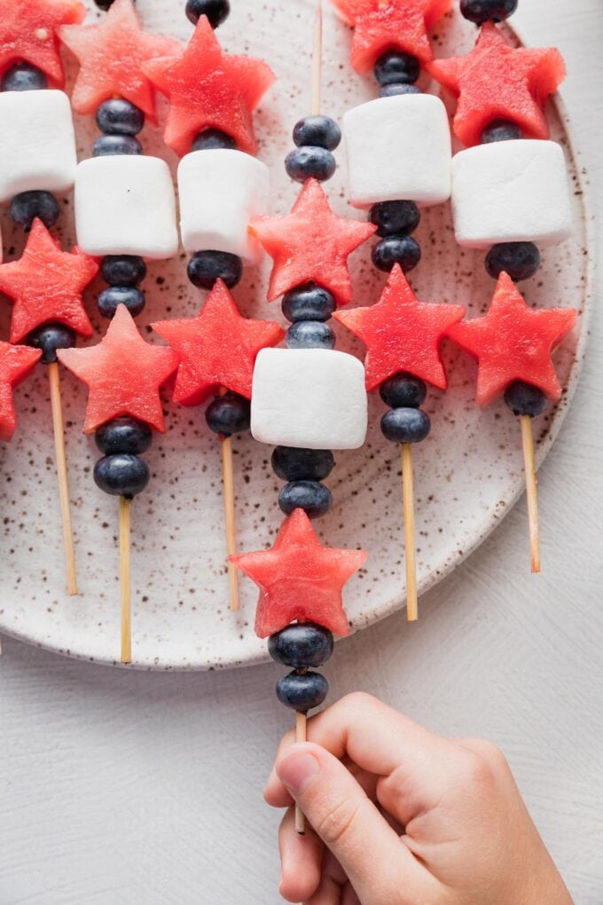 A child's hand picking up a bamboo skewer holding star-shaped watermelon cut-outs, fresh blueberries, and a large white marshmallow.
