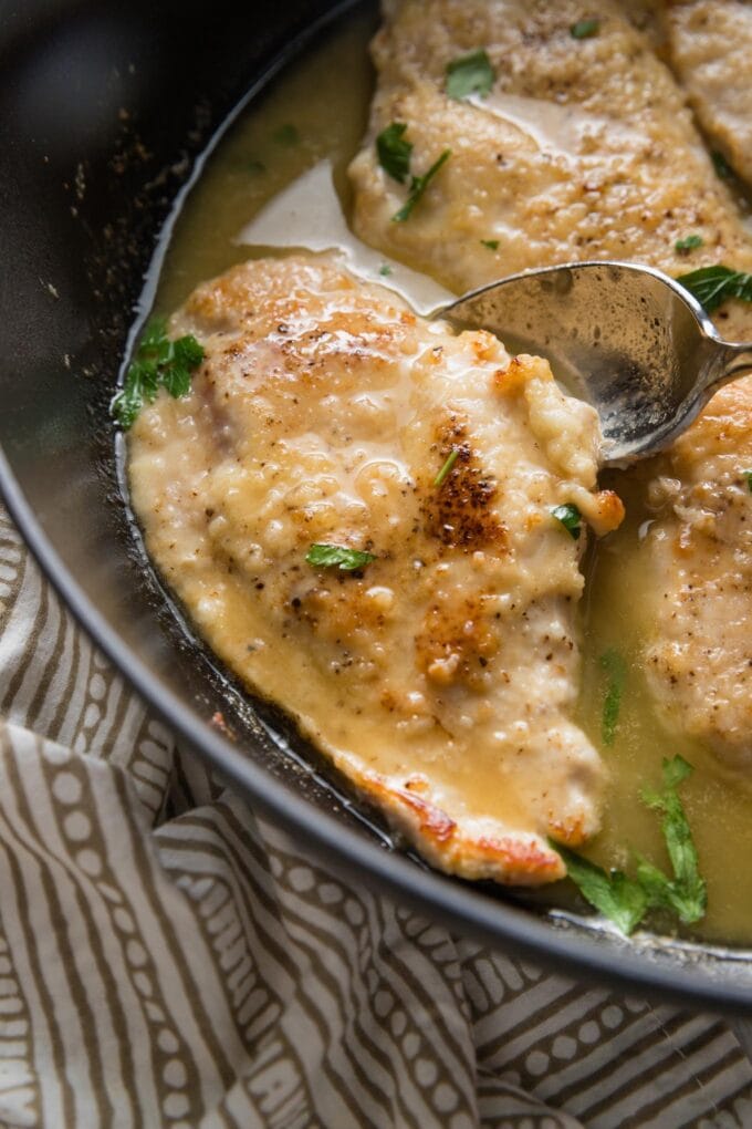 A spoon pouring a iight white wine sauce over chicken breasts in a skillet.