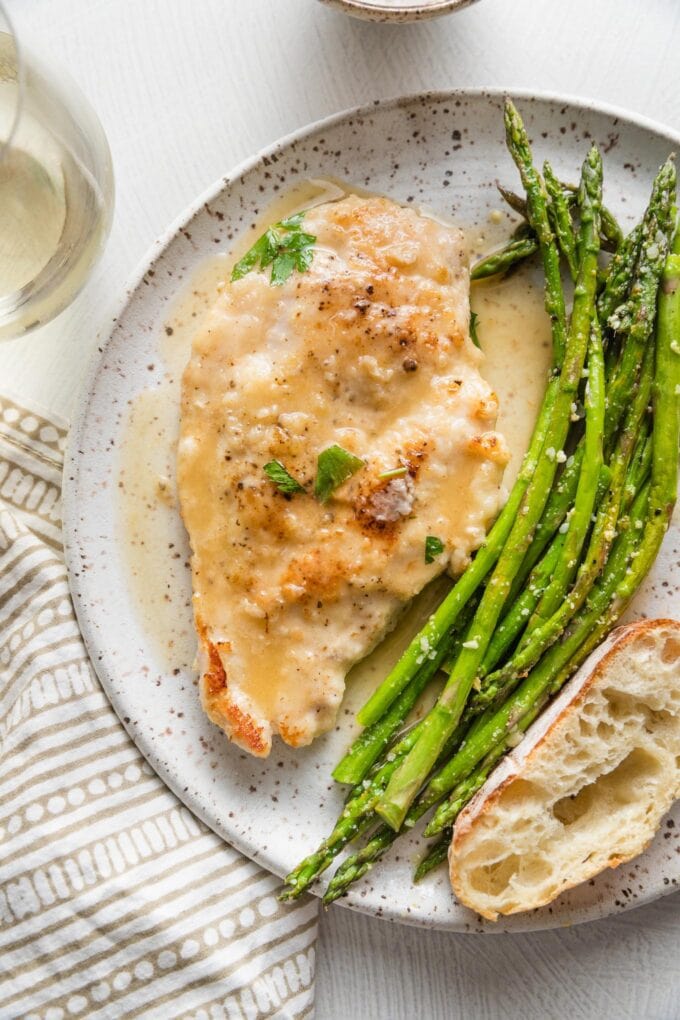 Small white plate with a tender pan-fried chicken breast in white wine sauce, roasted asparagus, and a slice of bread.