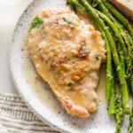 Small white plate with a tender pan-fried chicken breast in white wine sauce, roasted asparagus, and a slice of bread.