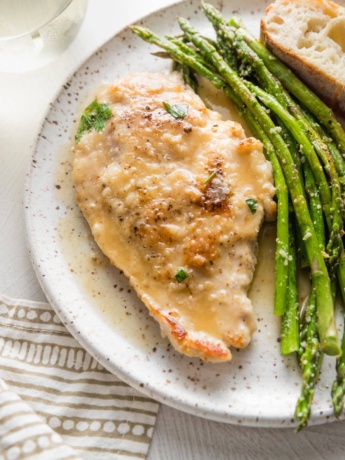 Small white plate with a tender pan-fried chicken breast in white wine sauce, roasted asparagus, and a slice of bread.