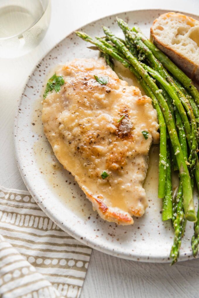 Small white plate with a tender pan-fried chicken breast in white wine sauce, roasted asparagus, and a slice of bread.