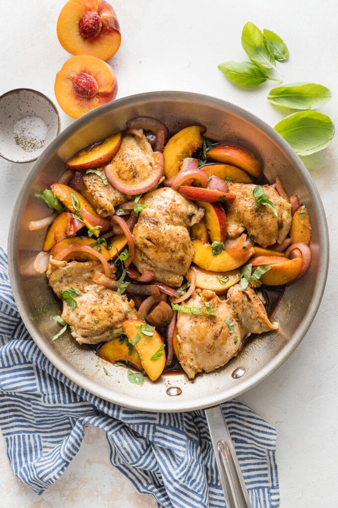 Overhead view of skillet with balsamic peach chicken surrounded by extra basil leaves, salt, and pepper.