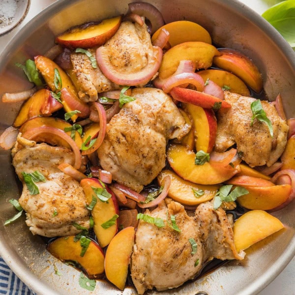 Large stainless steel skillet filled with a one pan dish of chicken thighs with sliced peaches, fresh basil, red onion, and a light balsamic glaze.