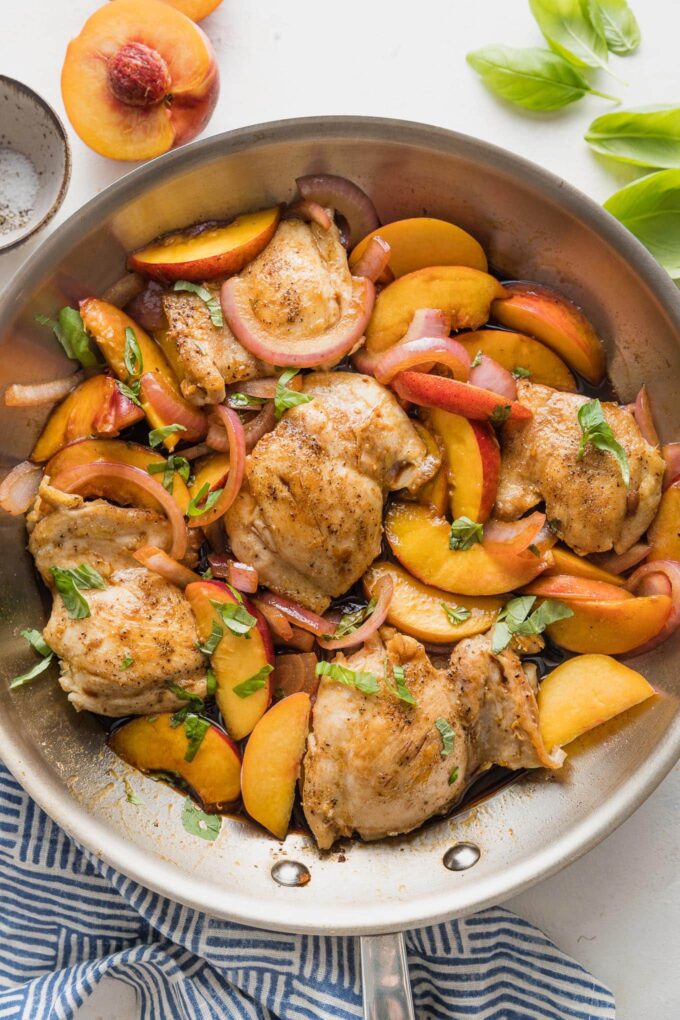 Large stainless steel skillet filled with a one pan dish of chicken thighs with sliced peaches, fresh basil, red onion, and a light balsamic glaze.