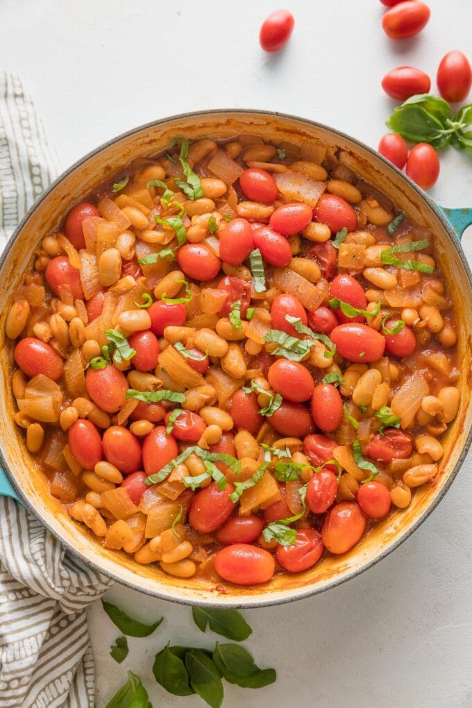 Overhead view of an enameled cast iron skillet filled with white beans and cherry tomatoes braised in a simple tomato balsamic vinegar sauce and garnished with slivers of fresh basil.