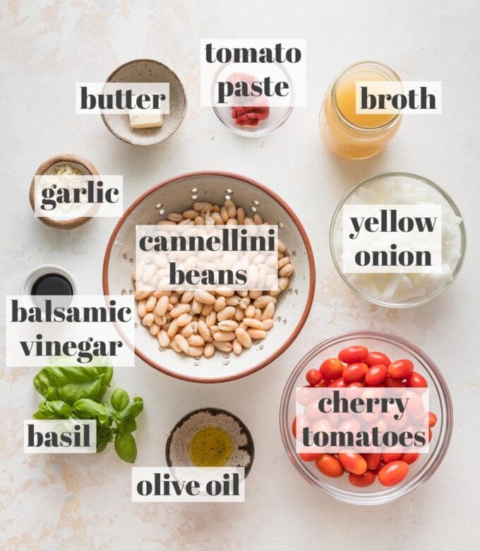 Labeled overhead photo of cannellini beans, cherry tomatoes, butter, tomato paste, vegetable broth, yellow onion, garlic, balsamic vinegar, fresh basil leaves, and olive oil measured and arranged in prep bowls.