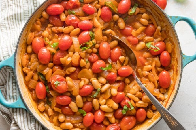 Close up of a spoon stirring braised white beans and tomatoes in a skillet.
