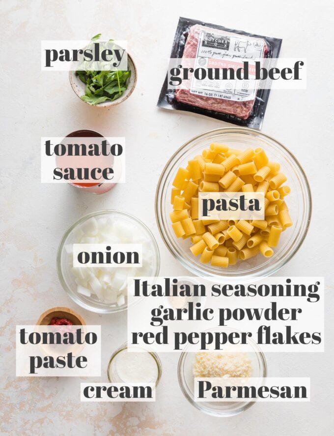 Labeled overhead image of ground beef in a package, dried pasta, chopped yellow onion, parsley, tomato sauce, tomato paste, cream, Parmesan, and spices all measured into prep bowls.