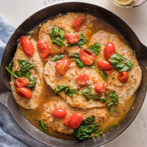 Cast iron skillet filled with chicken breasts in a light white wine sauce with spinach and tomatoes.