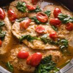 Close up of a cast iron skillet full of white wine chicken with baby spinach and burst cherry tomatoes.