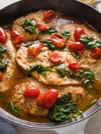 Close up of a cast iron skillet full of white wine chicken with baby spinach and burst cherry tomatoes.