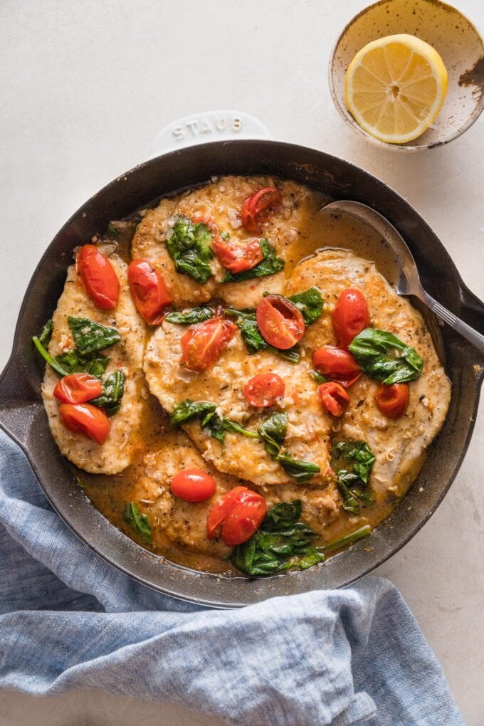 Overhead view of a spoon lifting sauce from a skillet filled with chicken breasts in a light white wine sauce with spinach and tomatoes.