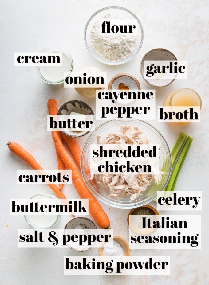 Labeled overhead image of carrots, celery, shredded chicken, chicken broth, cream, buttermilk, garlic, flour, a yellow onion, Italian seasoning, baking powder, salt, and pepper measured into prep bowls and ready to cook.