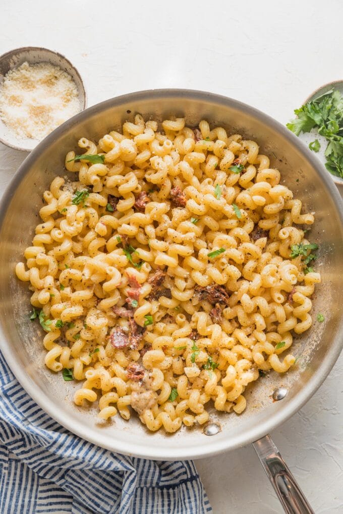 Wide-angle view of a stainless steel skillet full of creamy cavatappi pasta with extra Parmesan and parsley nearby for garnish.