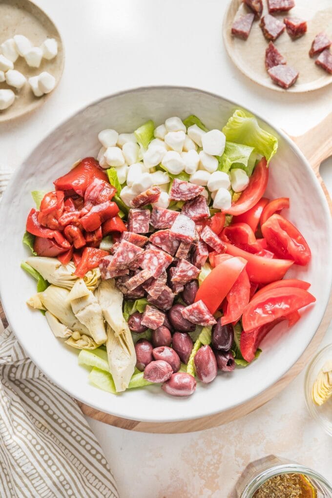 White bowl with lettuce, tomatoes, salami, and mozzarella added in sections.