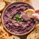 Close up of a crostini being dipped into a bowl of homemade olive tapenade.