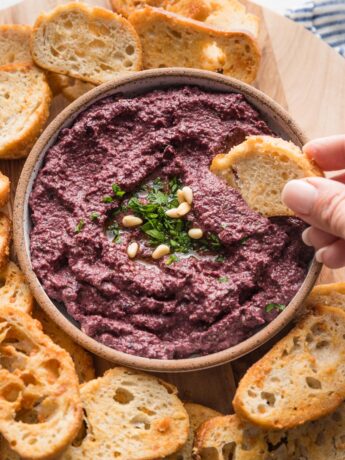 Close up of a crostini being dipped into a bowl of homemade olive tapenade.
