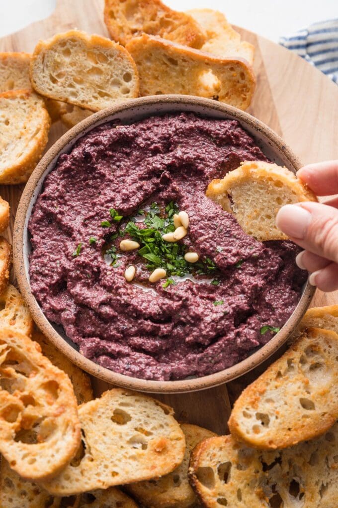 Close up of a crostini being dipped into a bowl of homemade olive tapenade.