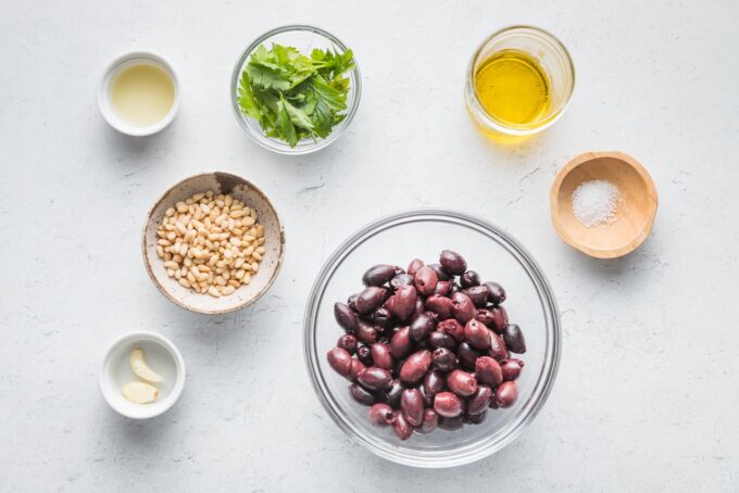 Overhead image of Kalamata olives, pine nuts, parsley, olive oil, lemon juice, garlic, and salt in prep bowls and ready to make into a Mediterranean dip.