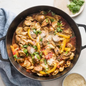 Deep cast iron skillet filled with tortellini, sausage, peppers, tomatoes, and Parmesan.