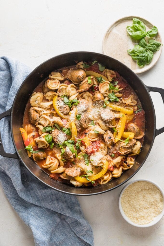 Deep cast iron skillet filled with tortellini, sausage, peppers, tomatoes, and Parmesan.