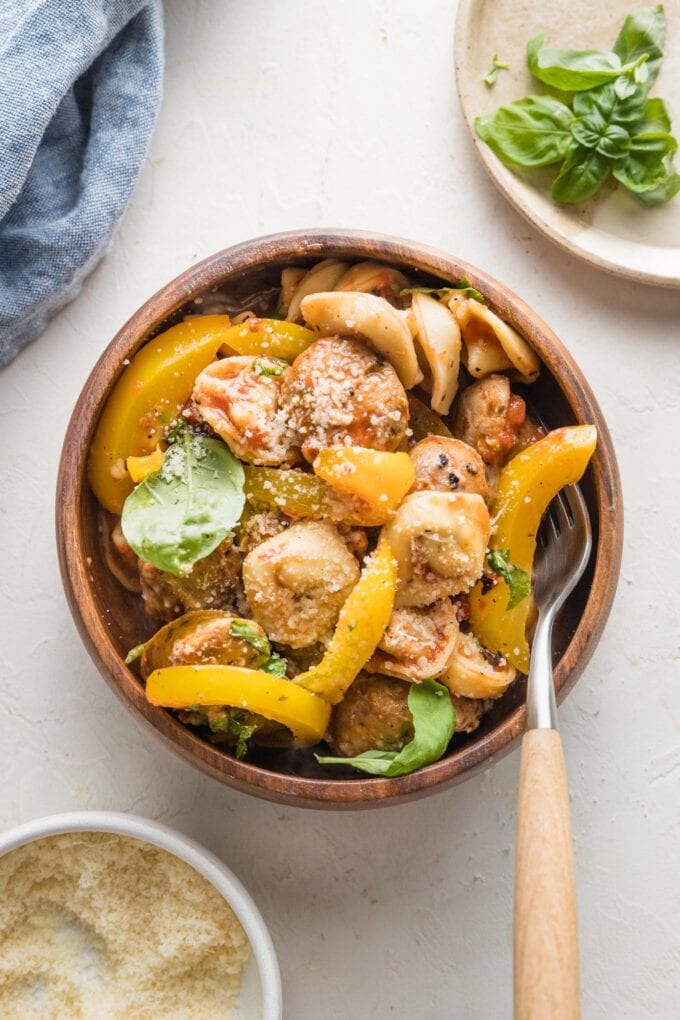 Small bowl with a cozy serving of tortellini cooked with sausage and peppers, garnished with Parmesan and fresh basil leaves.