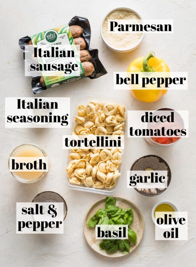 Labeled overhead image of refrigerated tortellini, pre-cooked Italian chicken sausage, a yellow bell pepper, canned diced tomatoes, broth, Parmesan, garlic cloves, olive oil, fresh basil leaves, and salt and pepper measured into prep bowls and ready to cook.
