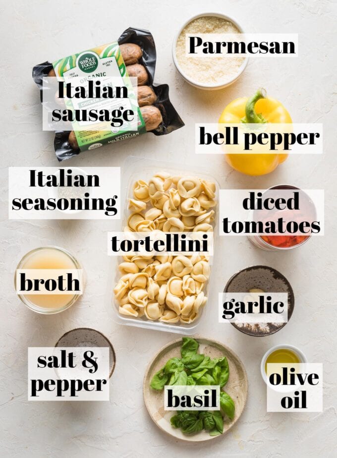 Labeled overhead image of refrigerated tortellini, pre-cooked Italian chicken sausage, a yellow bell pepper, canned diced tomatoes, broth, Parmesan, garlic cloves, olive oil, fresh basil leaves, and salt and pepper measured into prep bowls and ready to cook.