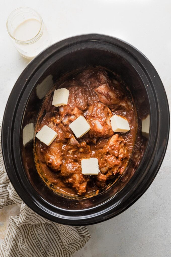 Large slow cooker filled with raw chicken, the ingredients for an Indian butter sauce, and pats of butter on top, ready to cook.