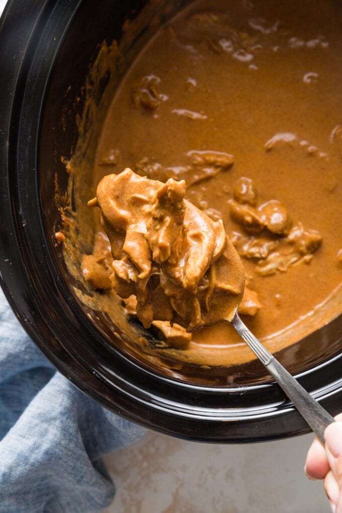 Serving spoon lifting a portion of cooked Indian butter chicken and sauce out of a large Crockpot.