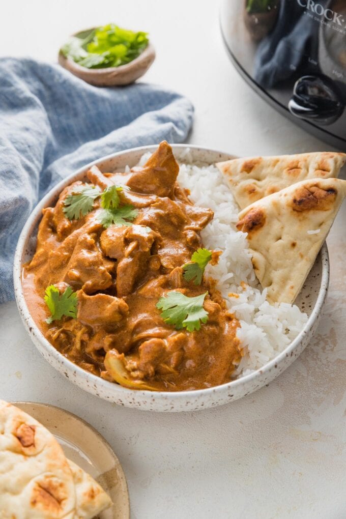 Bowl full of Indian butter chicken served with rice and naan, with a slow cooker in the background.
