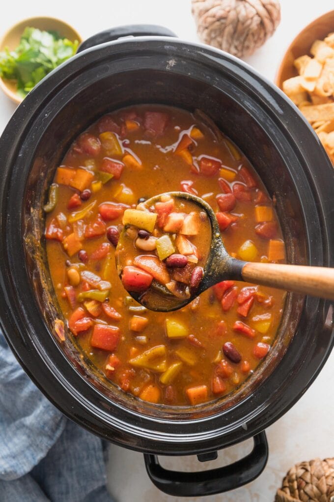 Overhead view of a ladle holding up a scoop of pumpkin chili directly above a big black slow cooker.